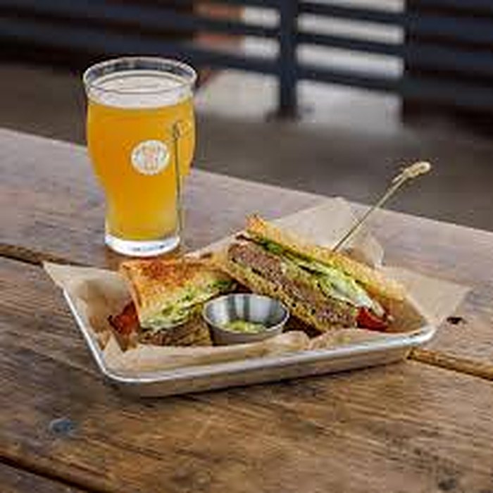 A pint of golden beer beside a toasted sandwich cut in half in a paper-lined tray on a wooden table.