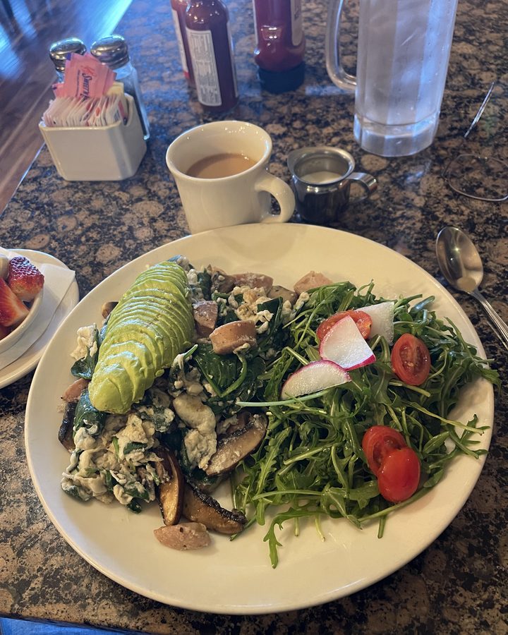 A breakfast plate of scrambled eggs, spinach, mushrooms, sausage and sliced avocado with an arugula and tomato salad, next to a cup of coffee.