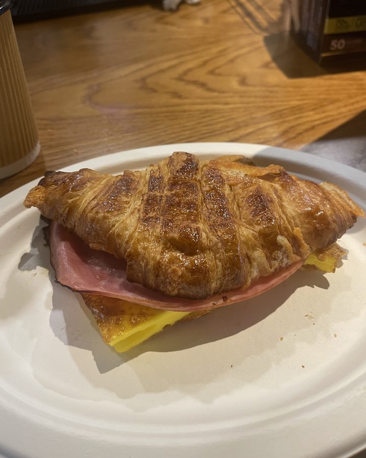 A grilled croissant sandwich with ham and egg on a paper plate at the counter.