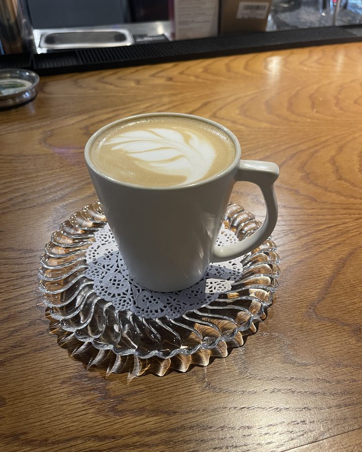 A latte with leaf-shaped foam art in a gray mug on a lace doily and a scalloped glass saucer, on a wooden bar.